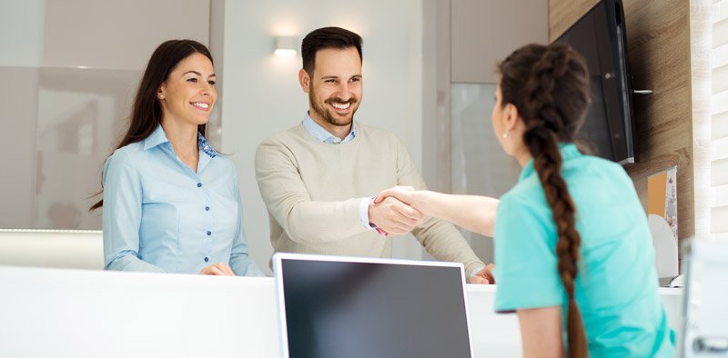 Patients greeting a dental clinic receptionist at the front desk during a visit.